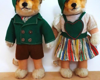 Puede incluir: Dos juguetes de peluche vintage con cara de zorro. Uno lleva una chaqueta verde y pantalones cortos marrones, el otro un vestido verde con un corazón y una falda a rayas. Ambos tienen gorras verdes y pies blancos y esponjosos. Están sobre una superficie beige.