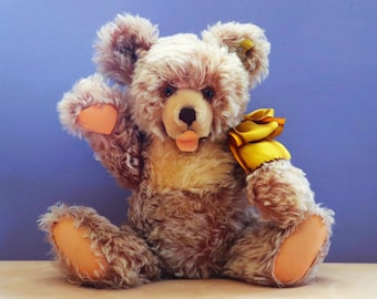 Pode incluir: Um urso de pelúcia marrom com peito amarelo e almofadas laranja. O urso tem uma fita amarela e está com a língua de fora. O fundo é azul sólido. O urso é feito de material de pelúcia.
