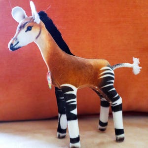 May include: A plush okapi toy with a brown body, black mane, and white and black striped legs. The toy has a white face, ears, and a small tail with a white tuft. The okapi is standing on a light-colored surface.
