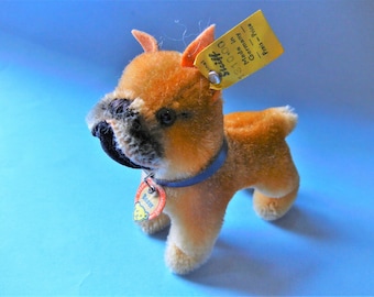 May include: A plush toy boxer dog with brown and tan fur, a black snout, and a blue collar. The dog has a yellow tag that reads "Steiff" and "Made in Germany". A small tag on the collar reads "Boxer".