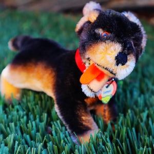 May include: A plush dog toy with black, tan, and cream fur, a red collar, and a tag. The dog's tongue is orange, and it is set against a backdrop of green grass.