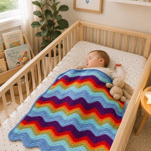 Peut inclure: Une couverture crochetée à la main présentant un motif chevron vibrant dans des tons de bleu clair, bleu foncé, vert, jaune, orange, rouge et violet. La couverture est posée sur un nourrisson endormi dans un berceau en bois avec un drap blanc à pois noirs.