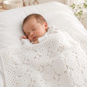 May include: A sleeping infant wearing a white long-sleeved shirt, covered by a white crocheted blanket featuring a repeating pattern of circular motifs and star-like shapes with a delicate openwork border along the edge of the textile.