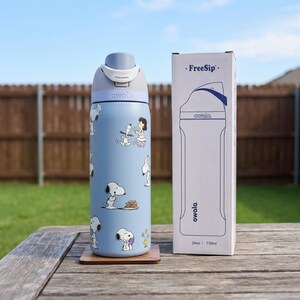 May include: A light blue Owala FreeSip water bottle with Snoopy and Woodstock graphics. The bottle is next to its packaging, which also features the Owala logo and a bottle outline. The bottle holds 24oz or 710ml.