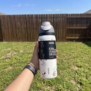 May include: A white Owala water bottle with a gray lid and black accents. The bottle features Snoopy illustrations and text that reads "Hi there. I'm leak-proof, I'm insulated, and I hoped built-in-ing." The bottle is held in front of a wooden fence and green grass.