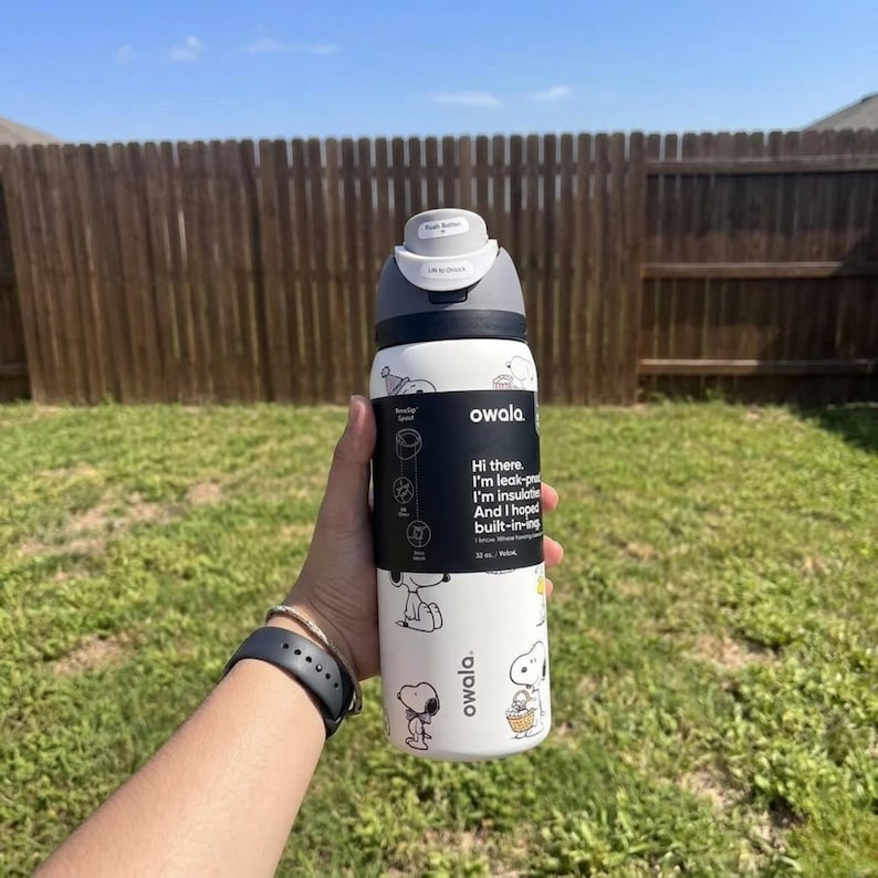 May include: A white Owala water bottle with a gray lid and black accents. The bottle features Snoopy illustrations and text that reads "Hi there. I'm leak-proof, I'm insulated, and I hoped built-in-ing." The bottle is held in front of a wooden fence and green grass.