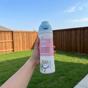 May include: A white and light blue Owala water bottle with a built-in straw. The bottle has a pink band with text that reads "Hi there. I'm leak proof. I'm insulated. And I have a built-in straw." A cartoon character is on the bottom.