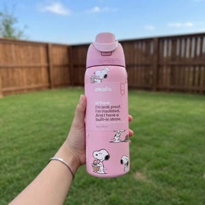 May include: Pink Owala water bottle with Snoopy designs. The bottle includes a built-in straw and text: "Hi there. I'm leak proof. I'm insulated. And I have a built-in straw." The bottle is 473 ml.