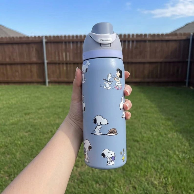 May include: A light blue water bottle featuring Snoopy and other Peanuts characters. The bottle has a gray lid with a push-button opening. The design includes Snoopy in various poses, with a few images of other characters. The bottle is held in front of a green lawn and a wooden fence.