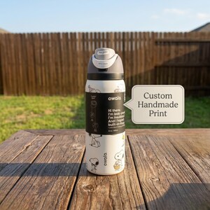May include: A white Owala insulated water bottle with a black and gray lid, featuring a Snoopy cartoon design. The bottle has a black band with text and is sitting on a wooden table. The text on the bottle says "Custom Handmade Print".