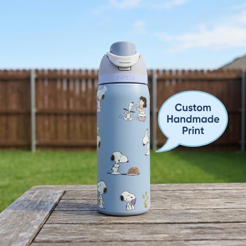 May include: A light blue water bottle with a gray lid and a white flip-top. The bottle is decorated with cartoon Snoopy characters. The text "Custom Handmade Print" is in a speech bubble. The brand name "awala" is printed on the bottle.