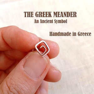 May include: A small silver-toned metal stud earring shaped like a square spiral, featuring the text THE GREEK MEANDER An Ancient Symbol Handmade in Greece. The earring has a metallic finish and a geometric design with a continuous line pattern.