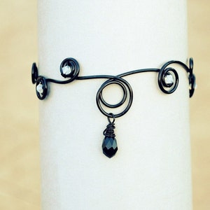 Peut inclure: Un bracelet en fil noir avec un cristal noir suspendu et des perles argentées. Le bracelet a un design délicat et complexe.