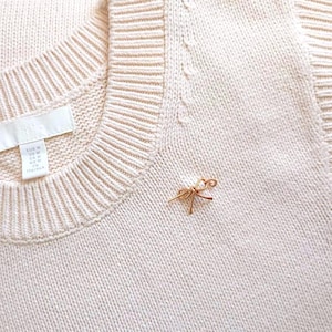 May include: A close-up of a cream-colored knit sweater with a small gold-colored bow pin. The sweater has a ribbed neckline and a tag on the inside. The bow pin is delicate and adds a touch of elegance to the garment.