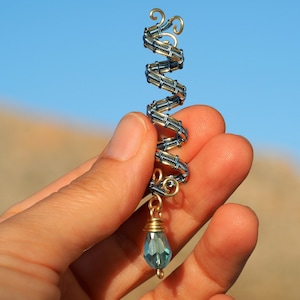 以下が含まれることがあります： ゴールドとブルーのワイヤーで巻かれた螺旋形のペンダント。下からブルーの涙型のクリスタルがぶら下がっている。