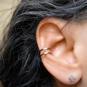 Pode incluir: Um ear cuff de banda dupla em tom de ouro rosa usado na cartilagem e um pequeno brinco de pino em forma de estrela prateada com pedras transparentes no lóbulo. As peças de joalharia são metálicas com um acabamento liso e polido no ear cuff.
