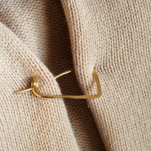 Op de afbeelding: Een goudkleurige metalen sjaalspeld is bevestigd aan een beige gebreide trui. De speld heeft een rechthoekige vorm met een kleine, ronde sluiting en een rechte speld. De gebreide textuur van de trui is zichtbaar.