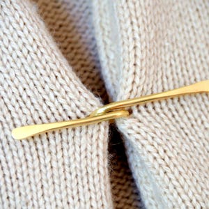 Könnte beinhalten: Gezeigt wird ein goldfarbener Metall-Pulloverclip, der die Vorderseite eines cremefarbenen Strickpullovers sichert. Der Clip hat ein schlichtes, elegantes Design mit einem gebogenen Haken und zwei flachen, länglichen Enden.