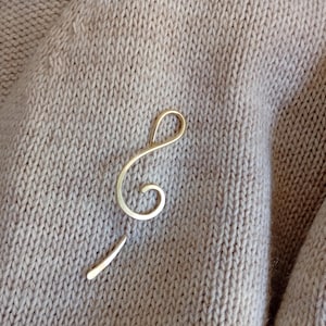 May include: A gold-colored treble clef brooch, a musical symbol, on a beige knitted fabric background.