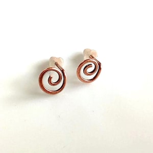 Puede incluir: Un par de pendientes de botón en espiral de color cobre. Cada pendiente presenta un diseño en espiral, con un cierre de poste. Los pendientes están sobre un fondo blanco, resaltando su apariencia intrincada y artesanal. Estos pendientes son un accesorio único.