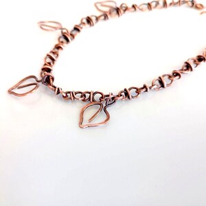 Op de afbeelding: Een koperdraad armband met een reeks onderling verbonden cirkelvormige schakels en meerdere bungelende bladvormige bedels. Het metaal heeft een warme, roodbruine tint met een getextureerde afwerking en een eenvoudige haaksluiting.