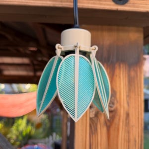 May include: A pendant light fixture featuring a cream-colored cylindrical base with a black power cord. Attached to the base are several teal-colored, leaf-shaped panels with a ribbed texture that surround the central light bulb area.