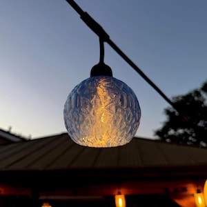 Puede incluir: Una lámpara exterior azul en forma de globo con una superficie texturizada. La luz cálida del interior del globo es visible. La lámpara cuelga de un cable negro contra el cielo crepuscular.