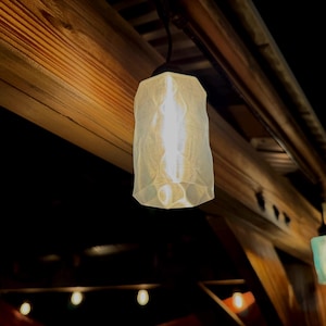May include: A hanging light fitting with a textured, off-white shade. The light is illuminated, casting a warm glow. The fitting is suspended from a dark cord and mounted on a wooden beam.