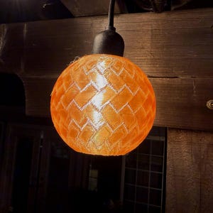 May include: A spherical orange light fitting with a woven design hangs from a black fitting. The light is on, emitting a warm glow. The fitting is suspended from a dark wooden beam.