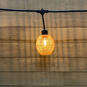 May include: A single, illuminated, spherical light bulb with a woven, cage-like design. The light is warm yellow, and the bulb hangs from a black wire against a wooden background. The light fixture is designed for outdoor use.