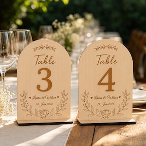 Può includere: Due segnatavolo in legno chiaro a forma di arco con la scritta Table 3 e Table 4. Ogni segnatavolo riporta i nomi Laura & Wilson, la data 14 Juin 2026 e motivi floreali incisi con foglie e rose. Poggiano su basi rettangolari in legno.