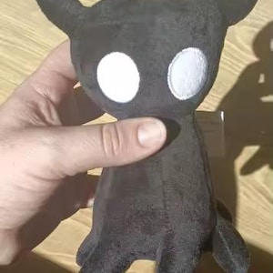 May include: A plush toy character featuring a solid black body with a soft, velvety texture. The figure has two prominent, curved horns extending from the top of its head and two large, circular, white embroidered eyes centered on its face.
