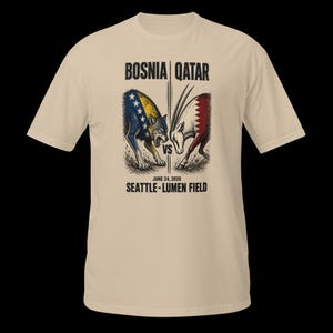 May include: A beige t-shirt with a graphic depicting a wolf and an oryx, symbolizing Bosnia and Qatar. The text "BOSNIA | QATAR" is above the image, with "VS" in the center. Below, it reads "JUNE 24, 2026 SEATTLE - LUMEN FIELD."