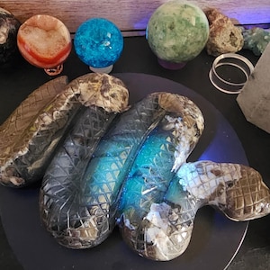 Puede incluir: Una escultura de serpiente enroscada tallada en piedra con un patrón de escamas texturizado en forma de cuadrícula. El cuerpo presenta una mezcla de tonos marrones y grises terrosos, con una sección azul translúcida vibrante en el centro.