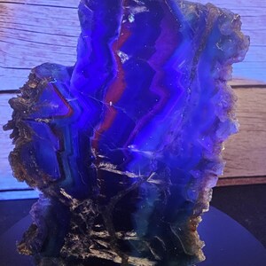 May include: A polished agate slice featuring vibrant bands of deep blue, purple, and reddish-brown hues. The translucent stone displays a jagged, natural crystalline edge with a smooth, flat face that reveals intricate, layered geometric patterns.