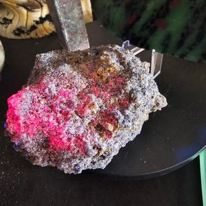 Puede incluir: Un espécimen mineral rugoso con una superficie texturizada que presenta parches vibrantes de rosa fluorescente, azul profundo y tonos dorados metálicos. La roca tiene una apariencia cristalina irregular y se exhibe sobre una base negra circular.