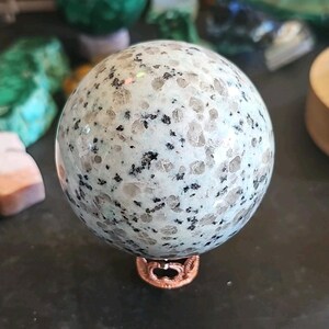 May include: A polished spherical stone featuring a light mint green base with scattered translucent crystalline inclusions and small black mineral flecks throughout the surface, resting on a small, ornate, metallic copper-colored circular stand.