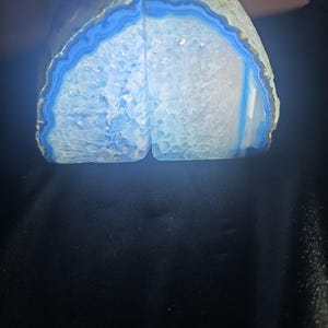 Puede incluir: Un par de sujetalibros de ágata azul con forma semicircular, un interior cristalino y bandas azules concéntricas en los bordes exteriores. Las piedras están iluminadas desde atrás, resaltando los cristales de cuarzo azul translúcidos.