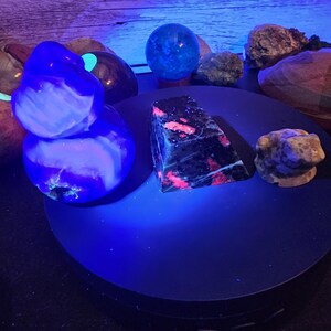 May include: A collection of mineral specimens under ultraviolet light, including a glowing blue and white rounded stone, a dark pyramid with fluorescent pink inclusions, and a small tan carved frog figure on a circular black display stand.