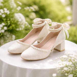 Könnte beinhalten: Ein Paar elfenbeinfarbene Schuhe mit niedrigem Absatz und Perlenakzenten. Die Schuhe haben eine runde Spitze, einen Knöchelriemen und eine dekorative Schleife. Die Absätze sind etwa 5 cm hoch. Die Schuhe stehen auf einer weißen Oberfläche.