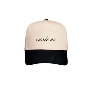 May include: A cream-coloured baseball cap with a black brim. The word "custom" is written in cursive across the front of the cap. The cap has a curved brim and a button on top.