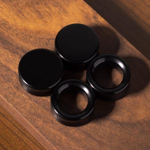 May include: Four black ear plugs arranged in a cluster. Two of the plugs are solid, smooth, circular discs, while the other two are hollow, ring-shaped tunnels with a polished, reflective finish, all featuring a consistent dark, glossy appearance.