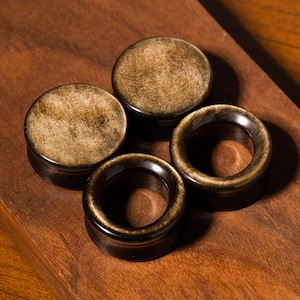 May include: Two circular ear plugs with a metallic gold and black finish. One plug is solid with a textured, shimmering surface, while the other is a hollow tunnel design. Both pieces feature a polished, reflective appearance with dark edges.