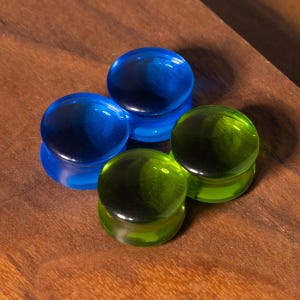 May include: Four circular ear plugs made of smooth, translucent material. Two of the plugs are a vibrant, deep blue color, while the other two are a bright, lime green. Each plug features a flat, polished front surface and a rounded edge profile.