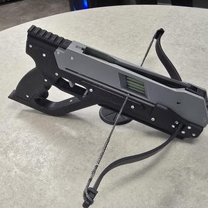 Könnte beinhalten: Schwarze und graue Armbrust mit strukturiertem Griff und grauem Körper. Die Bogensehne ist schwarz und weiß. Die Armbrust liegt auf einer hellgrauen Oberfläche.