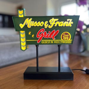 以下が含まれることがあります： Musso & Frank Grillの緑と黄色の電飾看板。テキストは「Oldest in Hollywood Since 1919」。看板は黒いベースとサポートポストを備えています。看板のテキストは黄色と赤です。