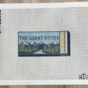 May include: Needlepoint canvas featuring a landscape scene with the text "THE GREAT DIVIDE". The design includes mountains, trees, and a body of water, rendered in shades of blue, green, and white. The canvas is on a white background with a grid pattern.
