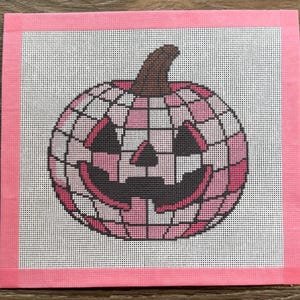 Könnte beinhalten: Ein quadratisches Stickbild mit einem Kürbis-Design im Disco-Kugel-Stil in Rosa. Der Kürbis besteht aus einem Gitter aus hell- und dunkelrosa Quadraten mit einem braunen Stiel sowie dunkelgrauen dreieckigen Augen, Nase und Mund.
