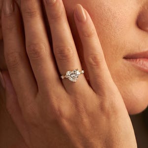May include: A gold ring featuring a prominent heart-shaped center stone flanked by two smaller round stones on each side. The band is thin and polished, showcasing a classic design that sits elegantly on the finger with a clear, faceted gemstone.