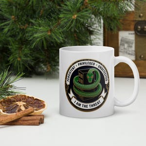 May include: White ceramic mug featuring a circular design with a green snake coiled around a green structure. The design includes the words "PERSISTENT", "PRIVILEGED", and "UNDETECTED". The text "I AM THE THREAT" is also present.